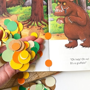 May include: A book with a colorful illustration of a brown monster with large teeth and horns. The book is open to a page with the text "Oh help! Oh no! It's a Gruffalo!"  The image also shows a hand holding a pile of colorful paper circles in green, orange, yellow, brown, and white.