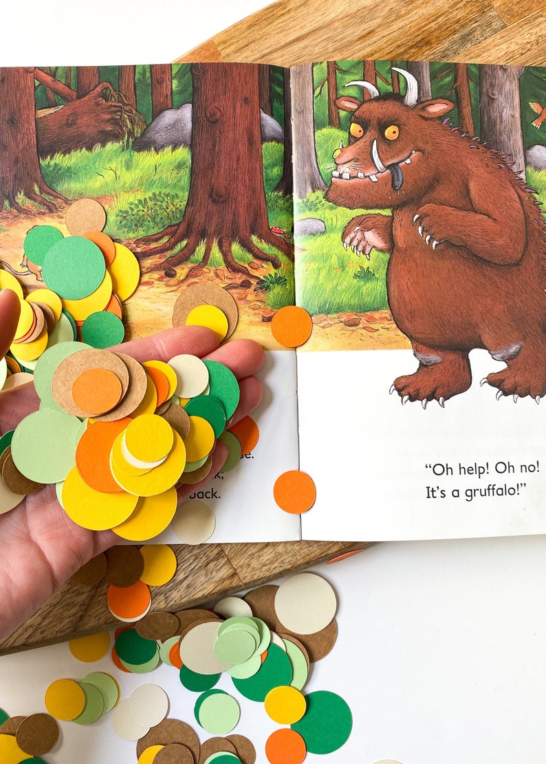 May include: A book with a colorful illustration of a brown monster with large teeth and horns. The book is open to a page with the text "Oh help! Oh no! It's a Gruffalo!"  The image also shows a hand holding a pile of colorful paper circles in green, orange, yellow, brown, and white.