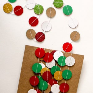 May include: A string of paper circles in red, green, white, gold, and brown with glitter accents. The circles are strung on white twine and are hanging on a brown paper background.