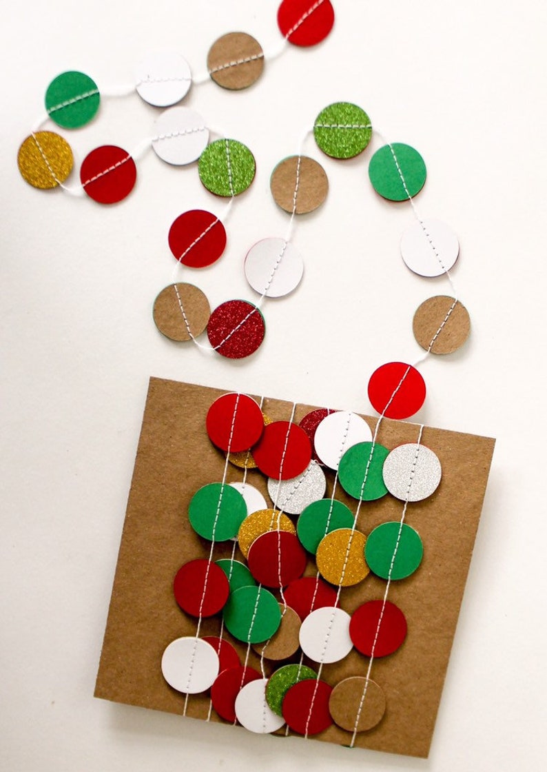 May include: A string of paper circles in red, green, white, gold, and brown with glitter accents. The circles are strung on white twine and are hanging on a brown paper background.