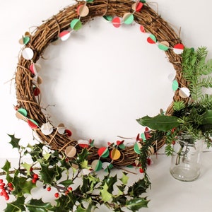 May include: A brown twig wreath decorated with a paper garland of red, green, white, and gold circles.  The wreath is accented with sprigs of holly and evergreen.