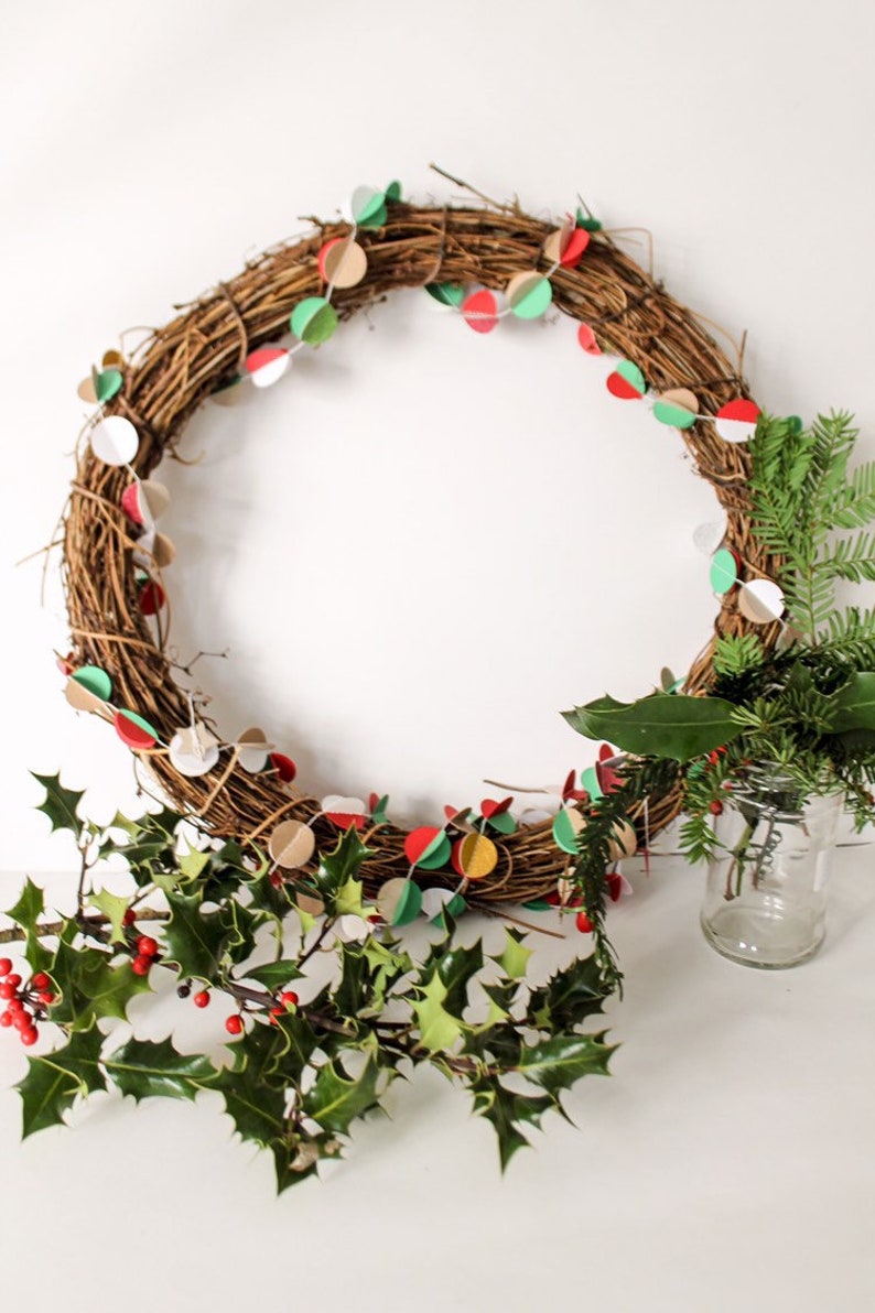 May include: A brown twig wreath decorated with a paper garland of red, green, white, and gold circles.  The wreath is accented with sprigs of holly and evergreen.