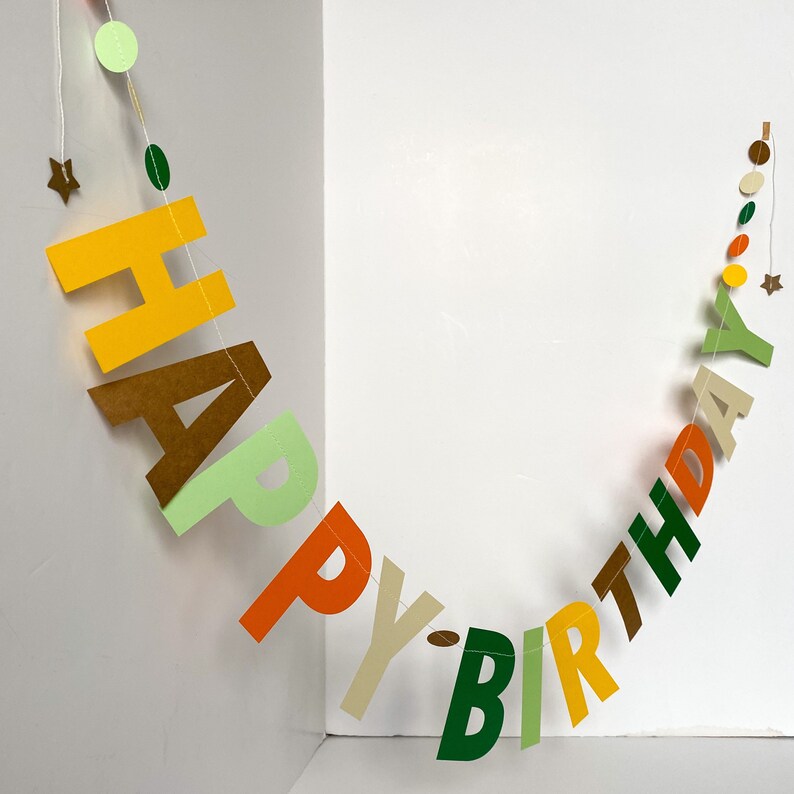 May include: A colorful paper banner with the words "Happy Birthday" in green, yellow, orange, brown, and white letters. The banner is strung together with small circles of different colors.