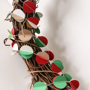 May include: A brown twig wreath decorated with a garland of paper circles in red, green, white, and brown. The garland is strung with white thread.