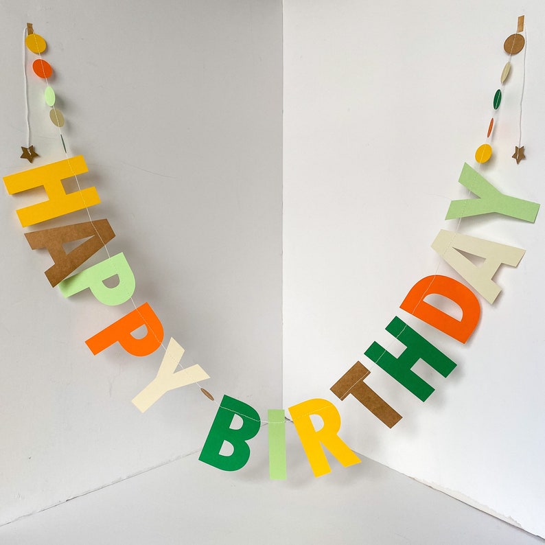 May include: A colorful paper banner with the words "Happy Birthday" in green, orange, yellow, brown, and cream letters. The banner is strung with a white string and decorated with small paper circles in various colors.