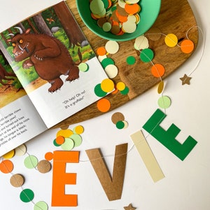 May include: A green, brown, and orange paper banner with the name "Evie" spelled out. The banner is strung with a white string and decorated with small paper circles in green, brown, orange, and cream. The background is white.