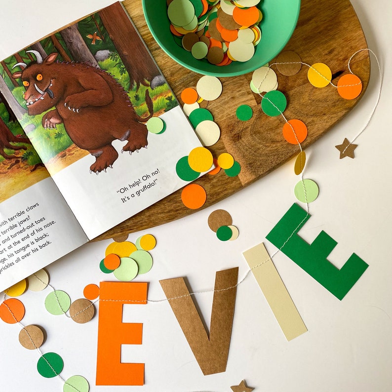 May include: A green, brown, and orange paper banner with the name "Evie" spelled out. The banner is strung with a white string and decorated with small paper circles in green, brown, orange, and cream. The background is white.