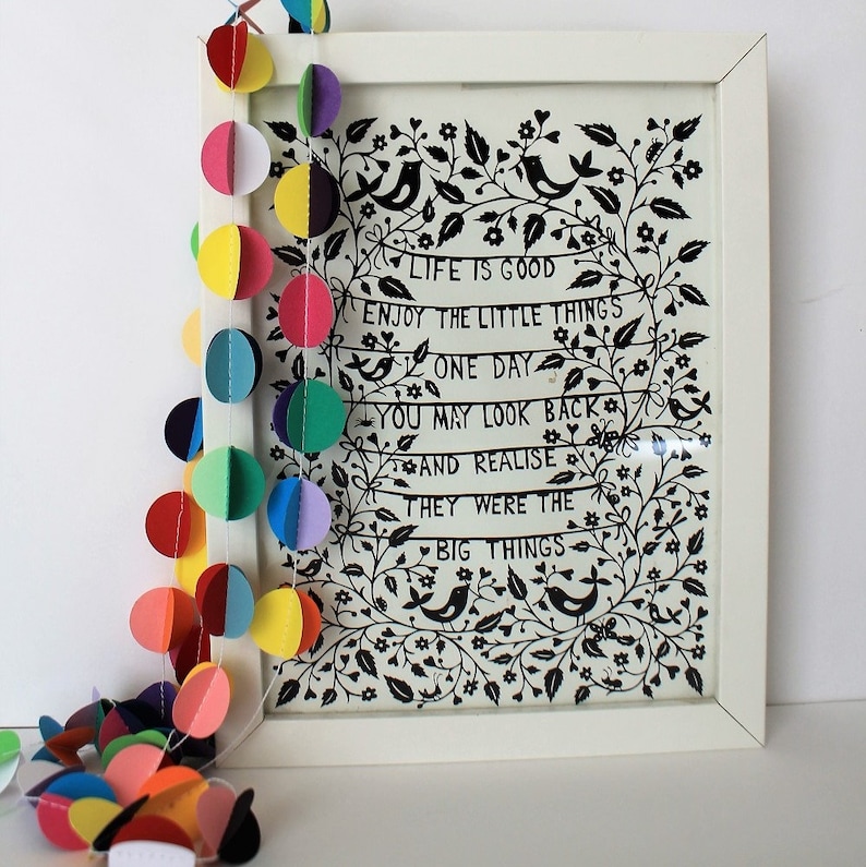 May include: A white framed print with a black and white illustration of a floral design and birds. The print has the text "LIFE IS GOOD ENJOY THE LITTLE THINGS ONE DAY YOU MAY LOOK BACK AND REALISE THEY WERE THE BIG THINGS". A colorful paper garland with circles hangs on the left side of the image.