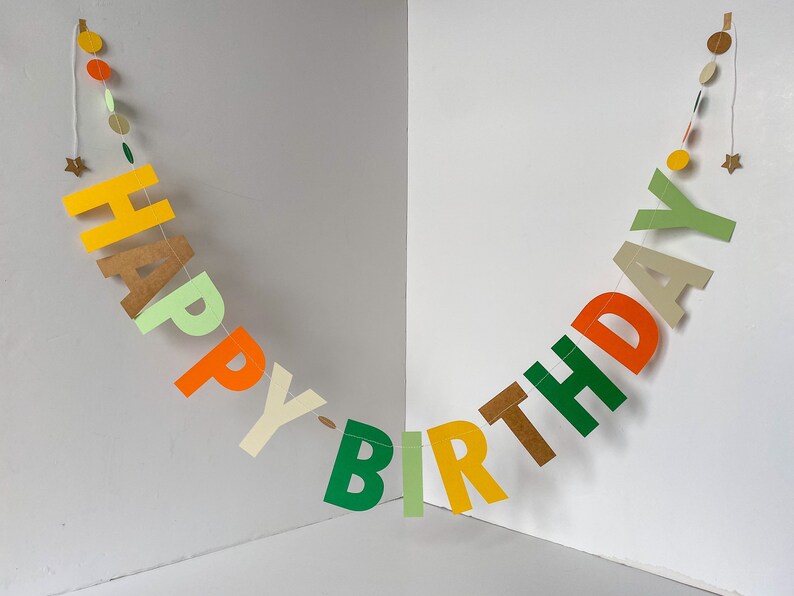 May include: A colorful paper banner with the words "Happy Birthday" in green, yellow, orange, and brown letters. The banner is strung with white thread and decorated with small circles in various colors.