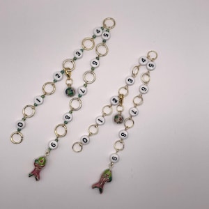 Könnte beinhalten: Zwei Armbänder mit Anhängern. Jedes Armband hat goldfarbene Kettenglieder, weiße Perlen mit schwarzen Zahlen und einen Fischanhänger. Die Armbänder haben auch dekorative Perlen und goldfarbene Ringe.