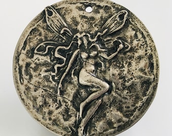 Può includere: Un ciondolo circolare in metallo color argento con un rilievo di una figura alata al centro. La superficie presenta una finitura testurizzata e usurata con ossidazione scura nelle aree incavate e un piccolo foro in alto per appenderlo.