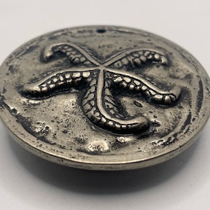 May include: A circular silver-toned metal button featuring a raised, textured starfish design in the centre. The surface of the button has a weathered, antiqued finish with dark recessed areas and a small hole at the top for attachment.