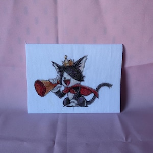 Könnte beinhalten: Kreuzstichbild einer Cartoon-Katze mit Krone und Umhang, die ein Megaphon hält. Die Katze ist schwarz-weiß mit rotem Umhang und Megaphon. Der Hintergrund ist ein hellrosa Stoff.