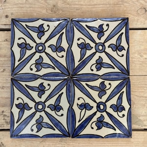 Op de afbeelding: Vier vierkante keramische tegels met een herhalend blauw-wit bloemmotief. Elke tegel heeft een centrale cirkel met stralende lijnen en gestileerde bladeren. De tegels zijn gerangschikt op een houten oppervlak.