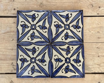 May include: Four square ceramic tiles with a repeating blue and white floral pattern. Each tile features a central circle with radiating lines and stylized leaves. The tiles are arranged on a wooden surface.
