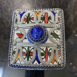 May include: A square, silver-trimmed box with a blue, red, green, and yellow geometric pattern. The box has a blue stone in the center and is decorated with silver filigree.