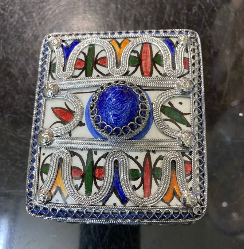 May include: A square, silver-trimmed box with a blue, red, green, and yellow geometric pattern. The box has a blue stone in the center and is decorated with silver filigree.