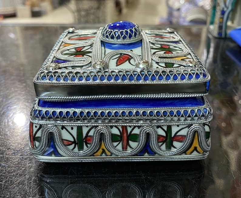 May include: A blue and white ceramic box with intricate silver filigree detailing. The box has a blue lid with a silver trim and a blue stone in the center.