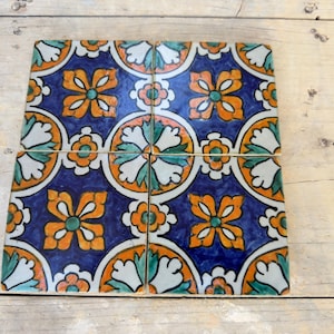 Op de afbeelding: Vier vierkante keramische tegels met een blauwe achtergrond en een herhalend patroon van oranje, witte en groene bloemen. De tegels zijn in een rasterpatroon gerangschikt.