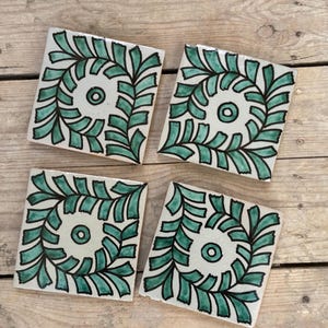 Puede incluir: Cuatro posavasos cuadrados de cerámica con fondo blanco y diseño de hojas verde azulado y negro. Cada posavasos presenta un círculo central y una espiral de hojas. Los posavasos están dispuestos sobre una superficie de madera.