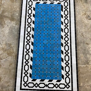 Op de afbeelding: Een blauw en wit Marokkaans tapijt met een geometrisch patroon. Het tapijt heeft een zwart-witte rand met een herhalend ontwerp.