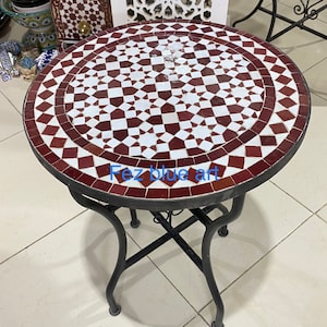 Op de afbeelding: Een ronde tafel met een blad van witte en bordeauxrode mozaïektegels en een zwart metalen onderstel. De tafel heeft een diameter van 61 cm en een hoogte van 71 cm. De tafel is perfect voor een kleine ruimte en voegt een vleugje Marokkaanse stijl toe aan elke kamer.