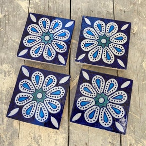 Può includere: Quattro piastrelle quadrate in ceramica con un design floreale blu e bianco. Ogni piastrella presenta un fiore stilizzato con petali blu e un centro bianco. Lo sfondo delle piastrelle è blu scuro.
