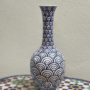 Peut inclure: Un vase élancé avec un motif de vagues blanc et bleu marine. Le vase a un col étroit et un corps arrondi. Le motif est répété symétriquement. Le vase est posé sur une table en mosaïque colorée.
