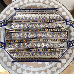 Peut inclure: Un plateau de service rectangulaire en c&eacute;ramique avec un motif g&eacute;om&eacute;trique d&eacute;taill&eacute; en bleu, marron, orange et vert sur fond blanc. Le plateau a des poign&eacute;es incurv&eacute;es et un bord bleu. Le plateau est pos&eacute; sur une table en mosa&iuml;que.