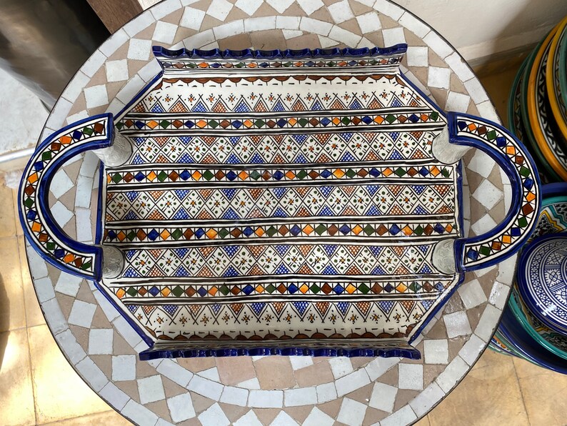 Peut inclure: Un plateau de service rectangulaire en c&eacute;ramique avec un motif g&eacute;om&eacute;trique d&eacute;taill&eacute; en bleu, marron, orange et vert sur fond blanc. Le plateau a des poign&eacute;es incurv&eacute;es et un bord bleu. Le plateau est pos&eacute; sur une table en mosa&iuml;que.