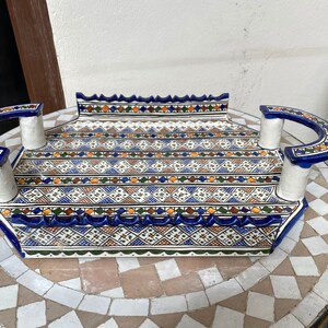 Peut inclure: Un plateau de service rectangulaire en c&eacute;ramique avec un motif g&eacute;om&eacute;trique d&eacute;taill&eacute; en bleu, blanc, orange et vert. Le plateau a des poign&eacute;es incurv&eacute;es et un bord festonn&eacute;. Le plateau est pos&eacute; sur une table en mosa&iuml;que.