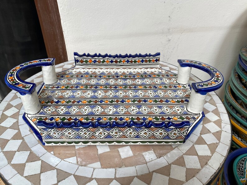 Peut inclure: Un plateau de service rectangulaire en c&eacute;ramique avec un motif g&eacute;om&eacute;trique d&eacute;taill&eacute; en bleu, blanc, orange et vert. Le plateau a des poign&eacute;es incurv&eacute;es et un bord festonn&eacute;. Le plateau est pos&eacute; sur une table en mosa&iuml;que.