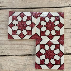 May include: Four square ceramic tiles with a geometric star pattern in burgundy and white. The tiles are arranged on a wooden surface, showcasing the decorative design. Each tile is approximately 15 cm square.