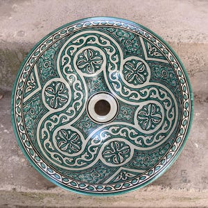 Può includere: Lavabo rotondo in ceramica con un motivo floreale ornato verde acqua e bianco. Il bacino presenta un foro di scarico centrale e un bordo decorativo. Il design include fiori stilizzati e linee a spirale.