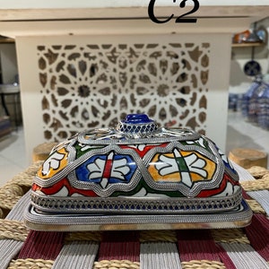 Peut inclure: Une boîte en céramique avec un couvercle en argent et un motif de carrelage marocain coloré. La boîte a un bijou bleu sur le dessus et le texte "C2" est visible en arrière-plan.