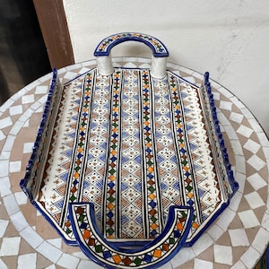 Peut inclure: Un plateau de service en c&eacute;ramique octogonal avec un motif g&eacute;om&eacute;trique bleu, orange, marron et vert. Le plateau a une base blanche, une bordure bleue et deux poign&eacute;es arqu&eacute;es. Il repose sur une table en mosa&iuml;que. Dimensions: environ 30 cm.