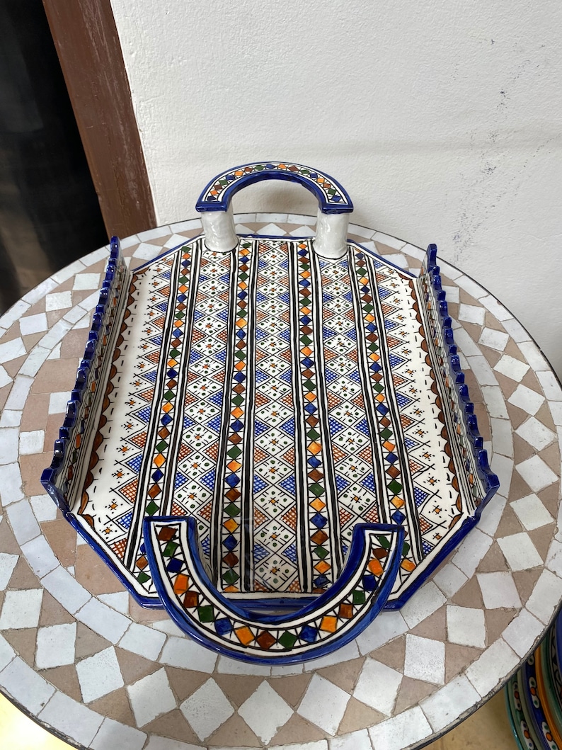 Peut inclure: Un plateau de service en c&eacute;ramique octogonal avec un motif g&eacute;om&eacute;trique bleu, orange, marron et vert. Le plateau a une base blanche, une bordure bleue et deux poign&eacute;es arqu&eacute;es. Il repose sur une table en mosa&iuml;que. Dimensions: environ 30 cm.