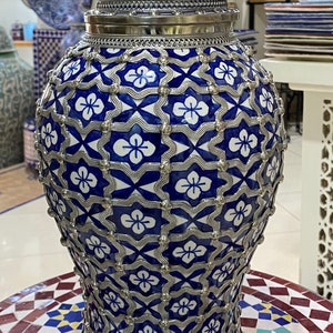 Puede incluir: Un jarr&oacute;n grande de cer&aacute;mica azul y blanco con intrincados detalles plateados. El jarr&oacute;n tiene un cuerpo redondo y una boca ancha y acampanada. El dise&ntilde;o presenta un patr&oacute;n repetitivo de flores y formas geom&eacute;tricas.