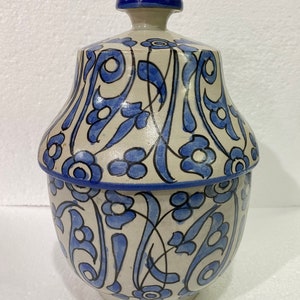 Puede incluir: Un tarro de cerámica blanca con tapa azul y diseños florales azules intrincados. El tarro tiene un cuerpo redondeado y una boca ancha.