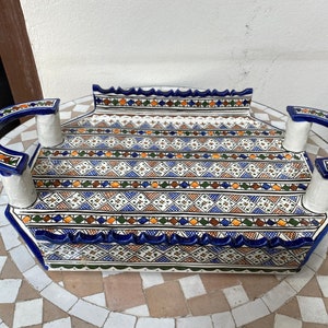 Peut inclure: Un plateau de service rectangulaire en c&eacute;ramique avec une base blanche et des motifs g&eacute;om&eacute;triques complexes en bleu, orange et vert. Le plateau comporte des poign&eacute;es incurv&eacute;es avec des d&eacute;tails bleus et blancs. Les bords sont festonn&eacute;s.