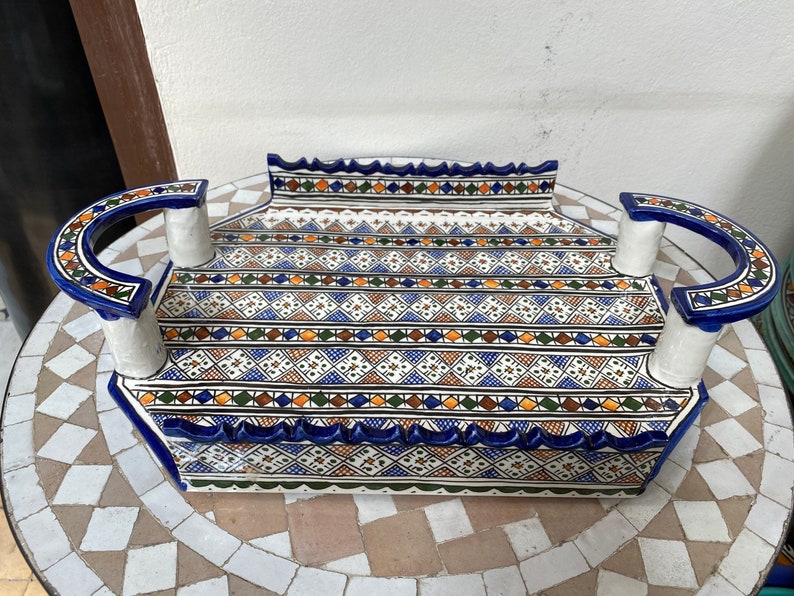 Peut inclure: Un plateau de service rectangulaire en c&eacute;ramique avec une base blanche et des motifs g&eacute;om&eacute;triques complexes en bleu, orange et vert. Le plateau comporte des poign&eacute;es incurv&eacute;es avec des d&eacute;tails bleus et blancs. Les bords sont festonn&eacute;s.