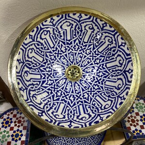 May include: Circular ceramic sink featuring a blue and white geometric design, framed by a gold-coloured rim. The sink includes a decorative drain cover, suitable for a bathroom or cloakroom.