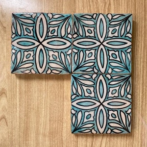 Puede incluir: Cuatro azulejos cuadrados de cerámica con un patrón geométrico azul y blanco. Los azulejos tienen un contorno negro y un fondo blanco con un centro azul. El patrón es un diseño repetitivo de hojas y óvalos.