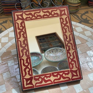 Puede incluir: Un espejo rectangular con un marco de azulejos rojo y crema. El marco tiene un patrón geométrico. El espejo refleja una pila de platos de cerámica de colores.