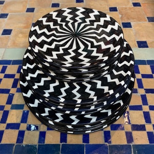 Op de afbeelding: Een stapel zwarte en witte keramische borden met een zigzagpatroon. De borden zijn spiraalsgewijs gestapeld, waardoor een optische illusie ontstaat.