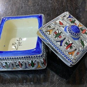 May include: A blue and white ceramic box with a silver filigree design. The box has a lid with a blue and silver design and a blue stone in the center. The box is inscribed with the word "FEZ".
