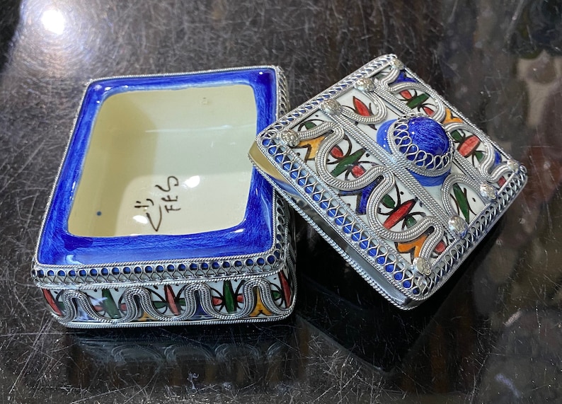 May include: A blue and white ceramic box with a silver filigree design. The box has a lid with a blue and silver design and a blue stone in the center. The box is inscribed with the word "FEZ".