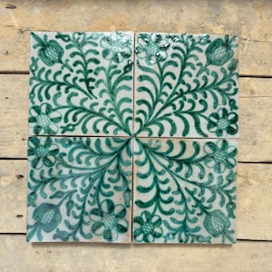 Puede incluir: Cuatro baldosas de cerámica cuadradas con fondo blanco y un patrón floral verde repetido. El diseño presenta flores y follaje estilizados, creando una disposición simétrica. Las baldosas están dispuestas juntas sobre una superficie de madera.