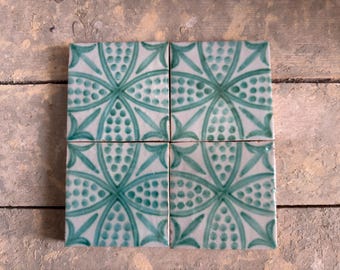 May include: Four square ceramic tiles with a white background and a teal floral design. Each tile features a symmetrical pattern of curved lines, dots, and leaf-like shapes. The tiles are arranged together, showcasing the repeating design.