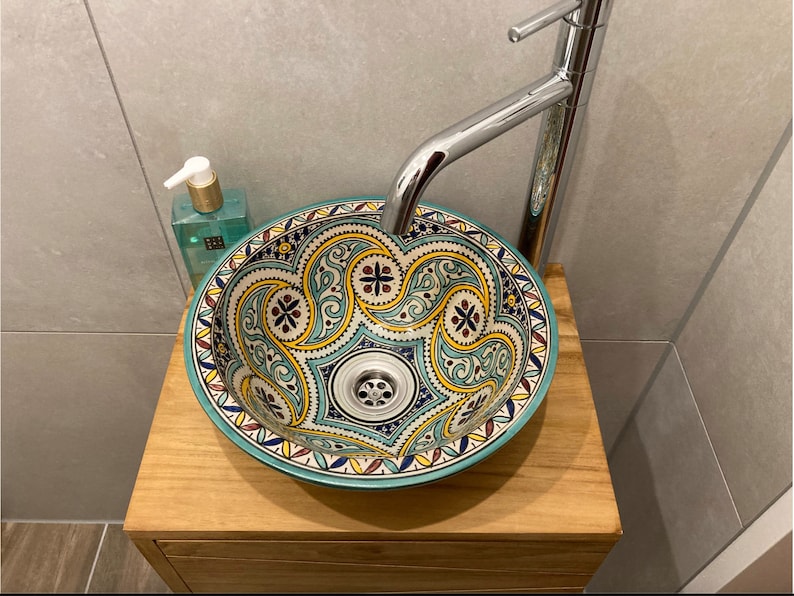 Puede incluir: Un lavabo de cer&aacute;mica turquesa y blanco con intrincados patrones florales amarillos y rojos. El lavabo est&aacute; montado en una encimera de madera con un grifo cromado.